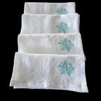 Four white cotton napkins stacked vertically on a dark reflective surface, each featuring a small turquoise embroidered coral motif near the top.