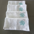 Four white linen napkins stacked vertically on a dark reflective surface, each featuring a small turquoise embroidered coral motif near the top.