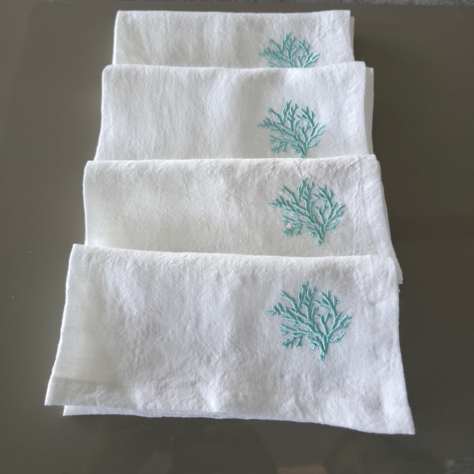 Four white linen napkins stacked vertically on a dark reflective surface, each featuring a small turquoise embroidered coral motif near the top.