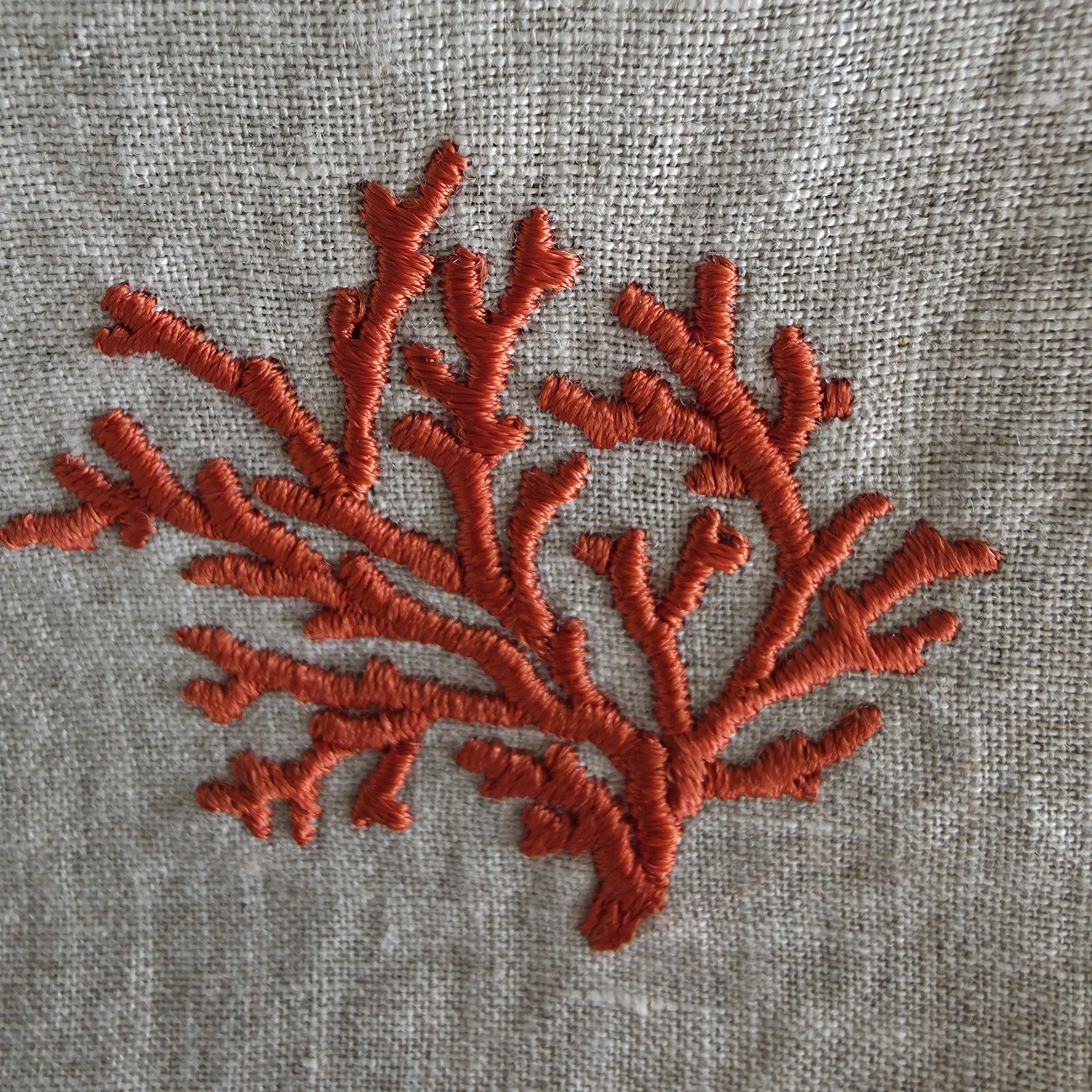 A close-up of coral-shaped embroidery in a rust orange-red thread on a textured light gray linen fabric background.