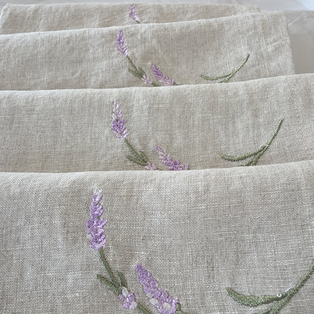 Close-up of folded beige linen fabric with embroidered lavender flowers and green stems. MI RE LA
