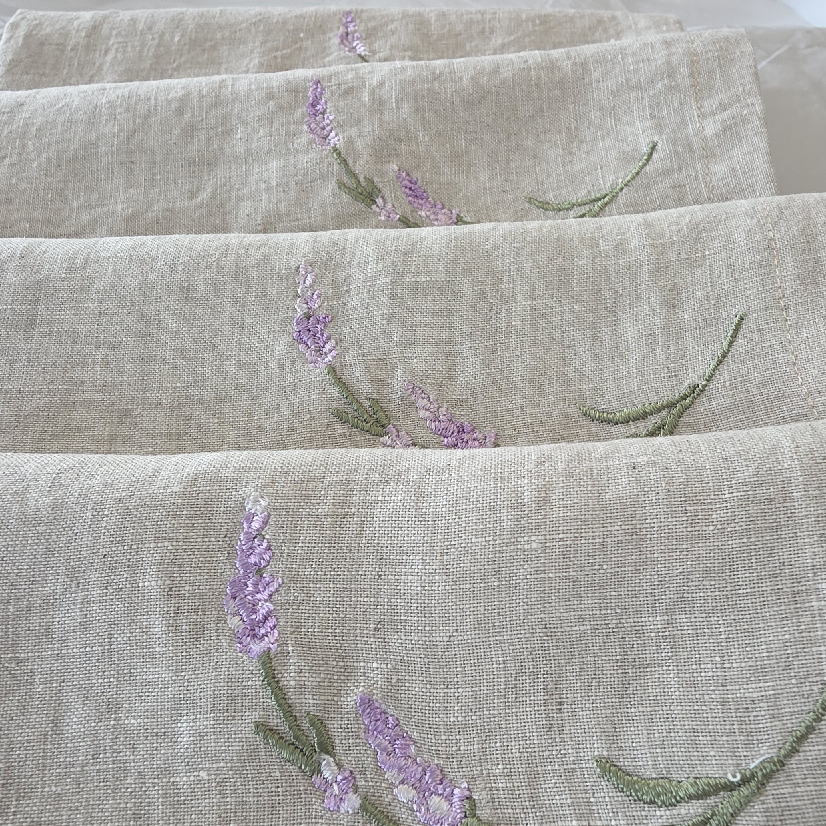 Close-up of folded beige linen fabric with embroidered lavender flowers and green stems. MI RE LA