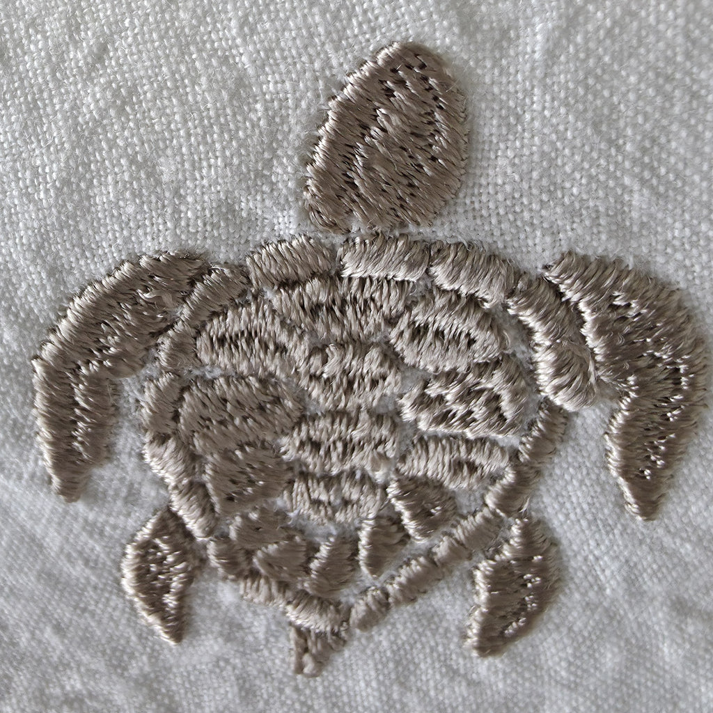 Close-up of a sea turtle embroidered design in taupe thread on a white textured fabric background. MI RE LA