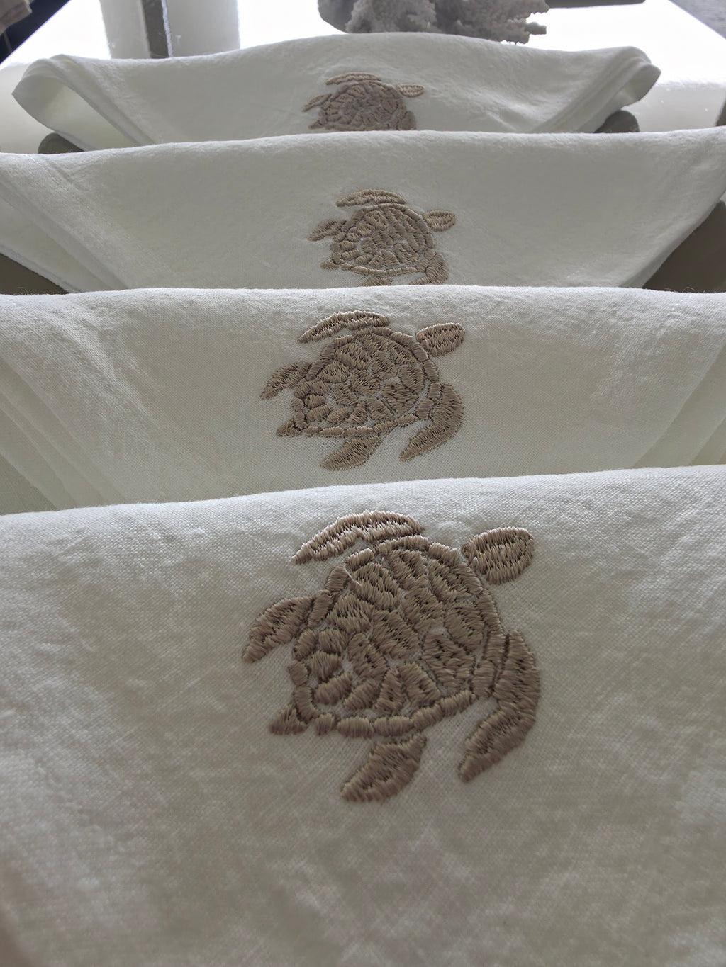 White towels with embroidered sea turtle motifs in a taupe-brown thread, shown hanging in a row. MI RE LA