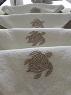 White towels with embroidered sea turtle motifs in a taupe-brown thread, shown hanging in a row. MI RE LA