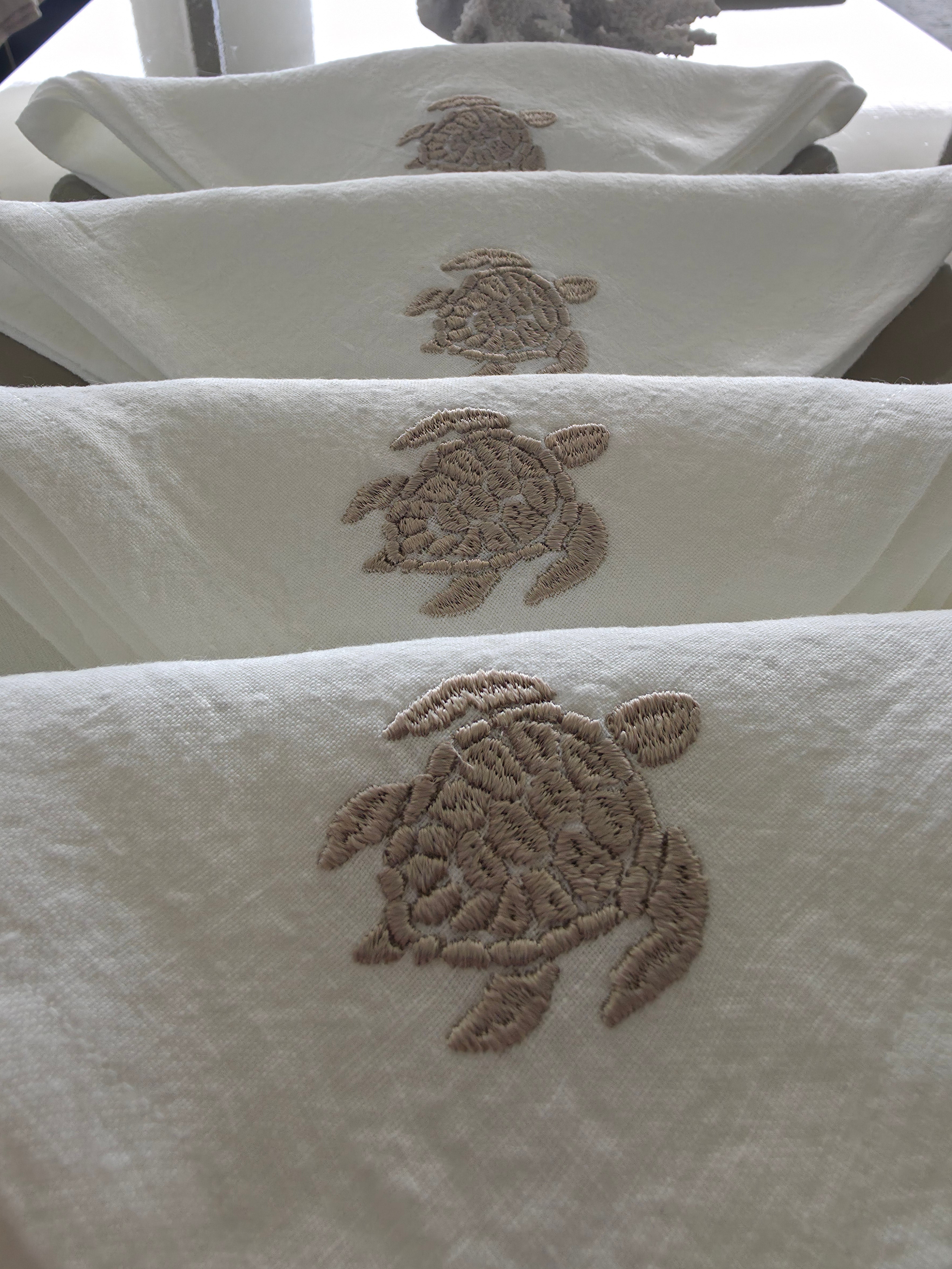 White towels with embroidered sea turtle motifs in a taupe-brown thread, shown hanging in a row. MI RE LA