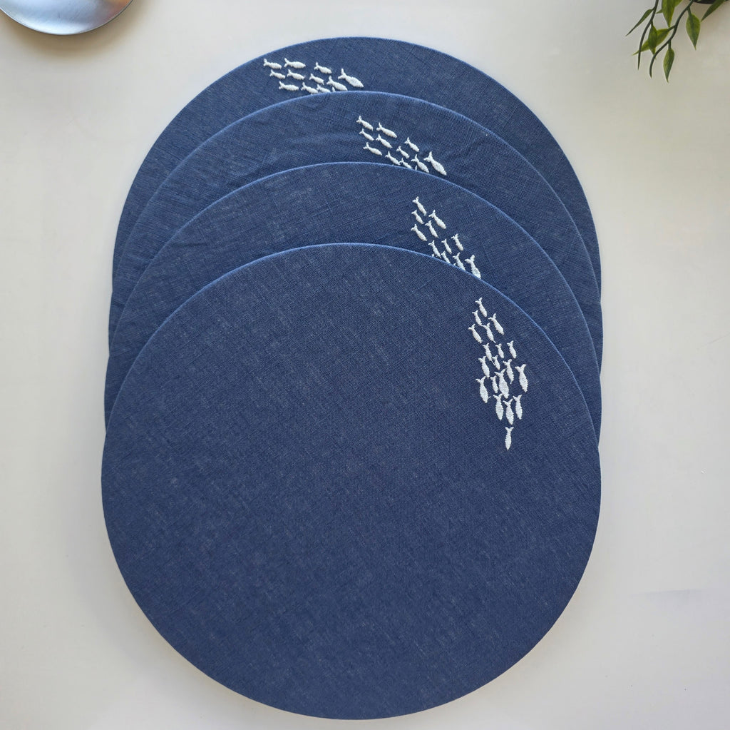 Four round navy blue placemats with white fish patterns, stacked on a light beige surface.