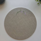 A round beige linen placemat with a delicate embroidered lavender stem and purple flower on the upper left, displayed on a clean white surface. MI RE LA