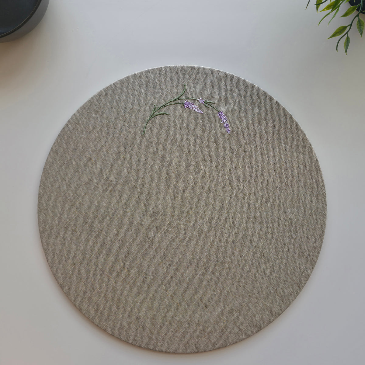 A round beige linen placemat with a delicate embroidered lavender stem and purple flower on the upper left, displayed on a clean white surface. MI RE LA