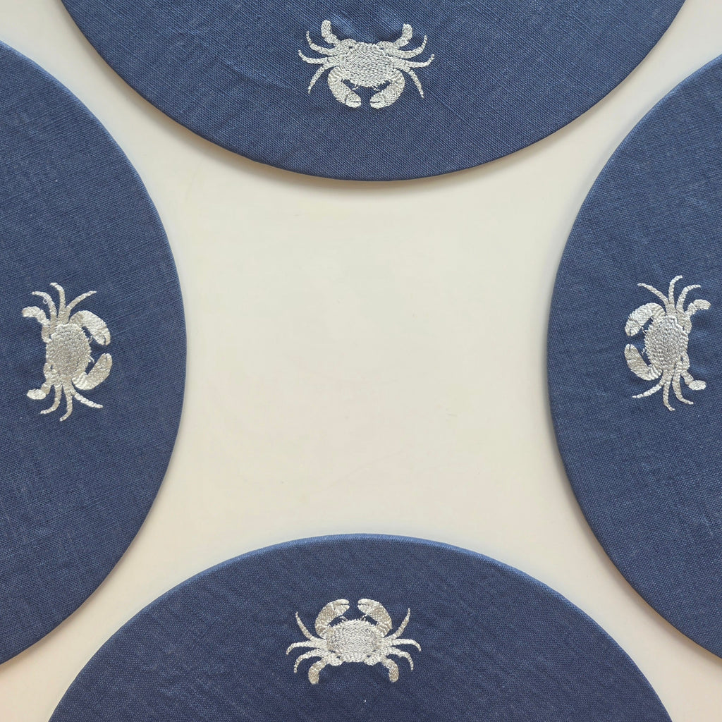 Four round navy blue coasters with white embroidered crabs, arranged in a square pattern on a light cream background.