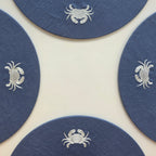 Four round navy blue coasters with white embroidered crabs, arranged in a square pattern on a light cream background.