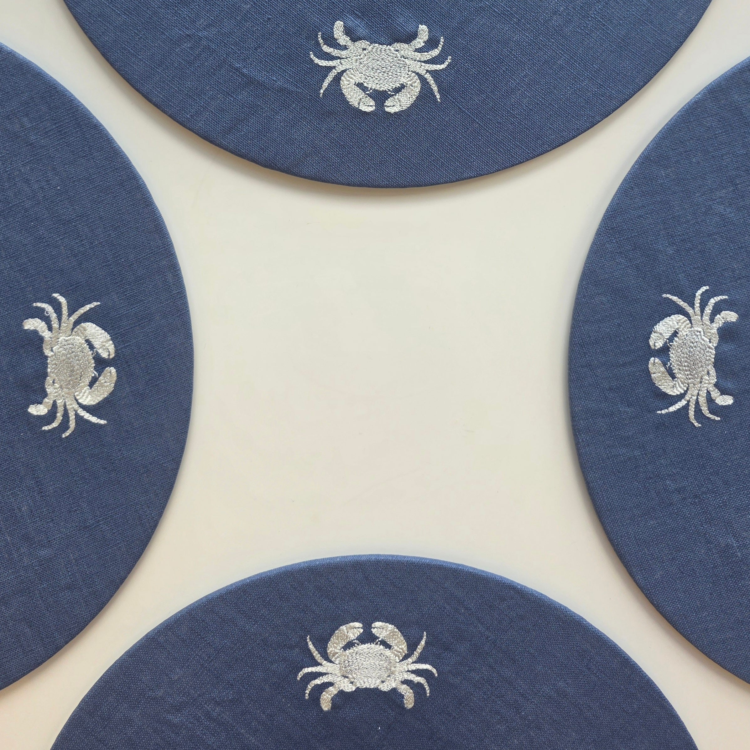 Four round navy blue coasters with white embroidered crabs, arranged in a square pattern on a light cream background.