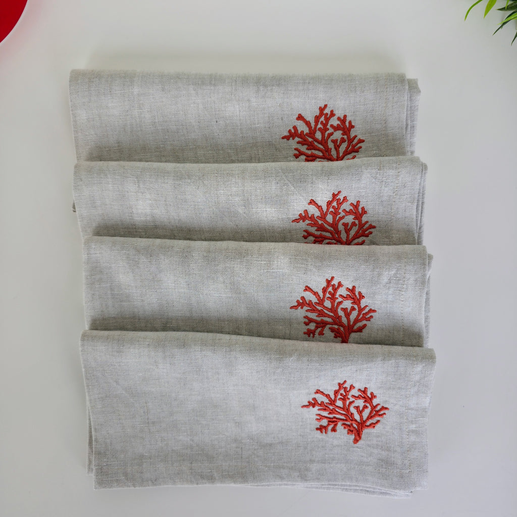 Four folded light gray linen napkins stacked on a white surface, each printed with a small red coral motif near the top.