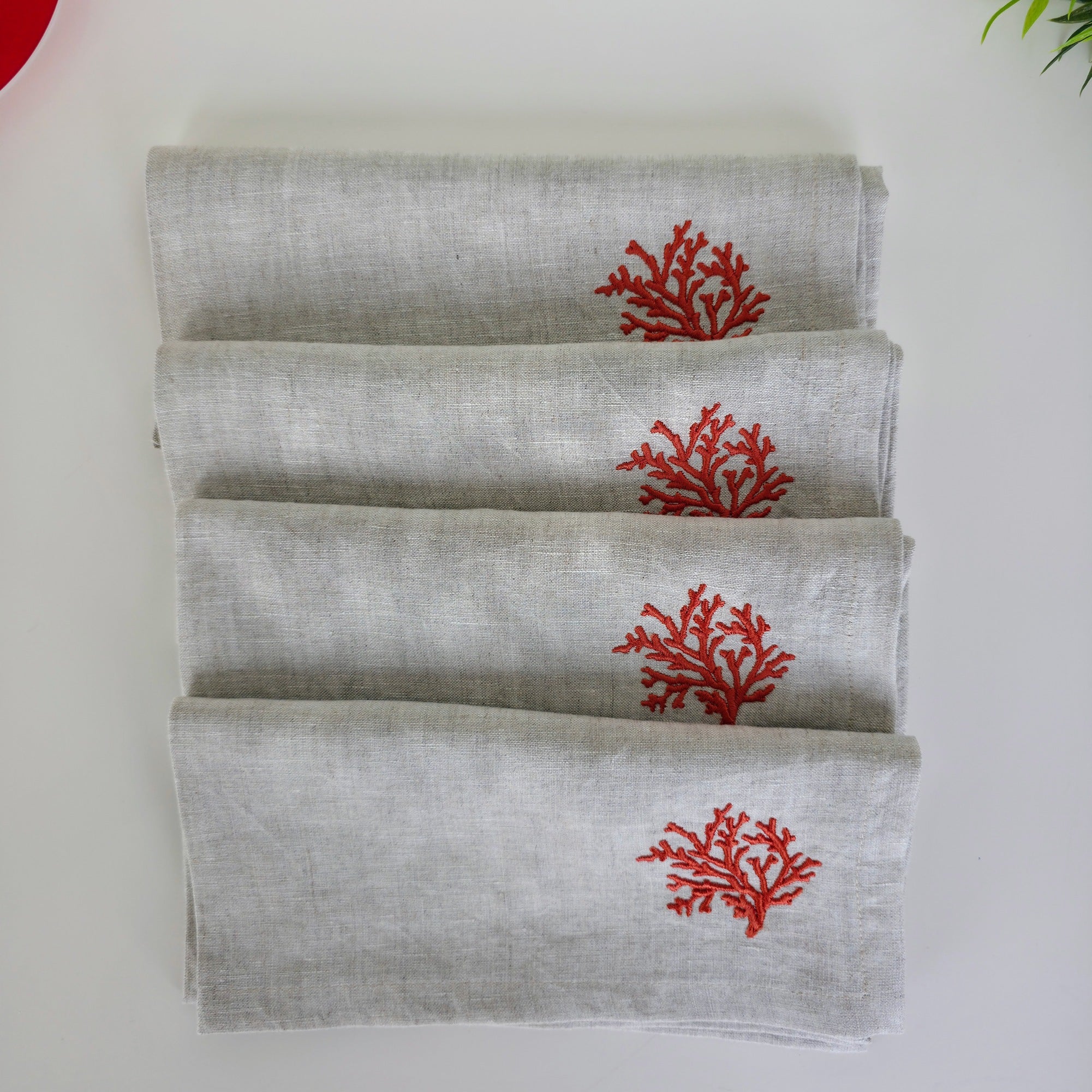Four folded light gray linen napkins stacked on a white surface, each printed with a small red coral motif near the top.