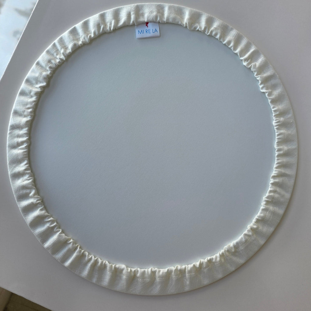 A circular white fabric-covered ring with a gathered, ruffled edge, photographed from above on a light gray surface.