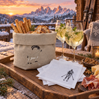 A rustic wooden tray set in a snowy mountain lodge at sunset, featuring a beige linen basket filled with breadsticks and bread, two filled wine glasses, white napkins with a black ski-and-snowboard print, and a charcuterie board with prosciutto and cheese.