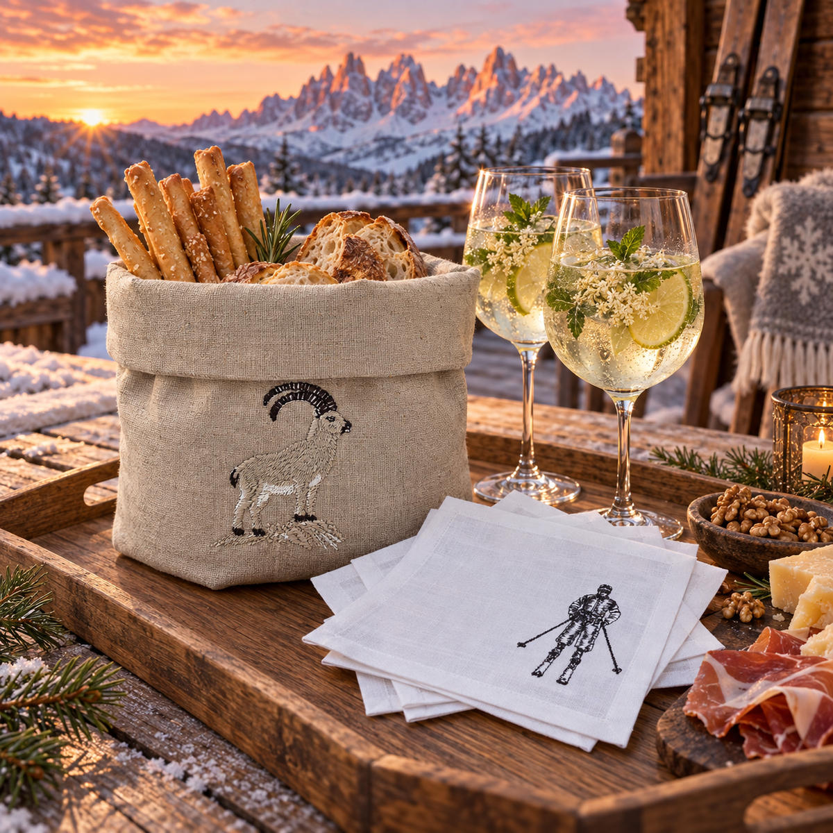 A rustic wooden tray set in a snowy mountain lodge at sunset, featuring a beige linen basket filled with breadsticks and bread, two filled wine glasses, white napkins with a black ski-and-snowboard print, and a charcuterie board with prosciutto and cheese.