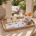 A wooden serving tray styled on a stone table in an outdoor patio setting, holding two clear glasses with lime slices, a glass carafe of water, white napkins with blue patterns, a white bread container with blue floral print, and a small bowl of nuts.