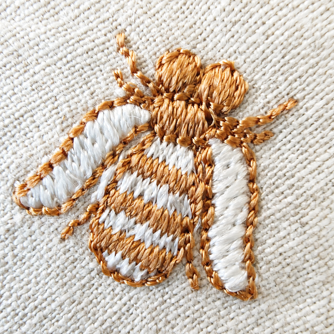 A close-up of a decorative embroidered bee applique on a textured cream fabric background. MI RE LA