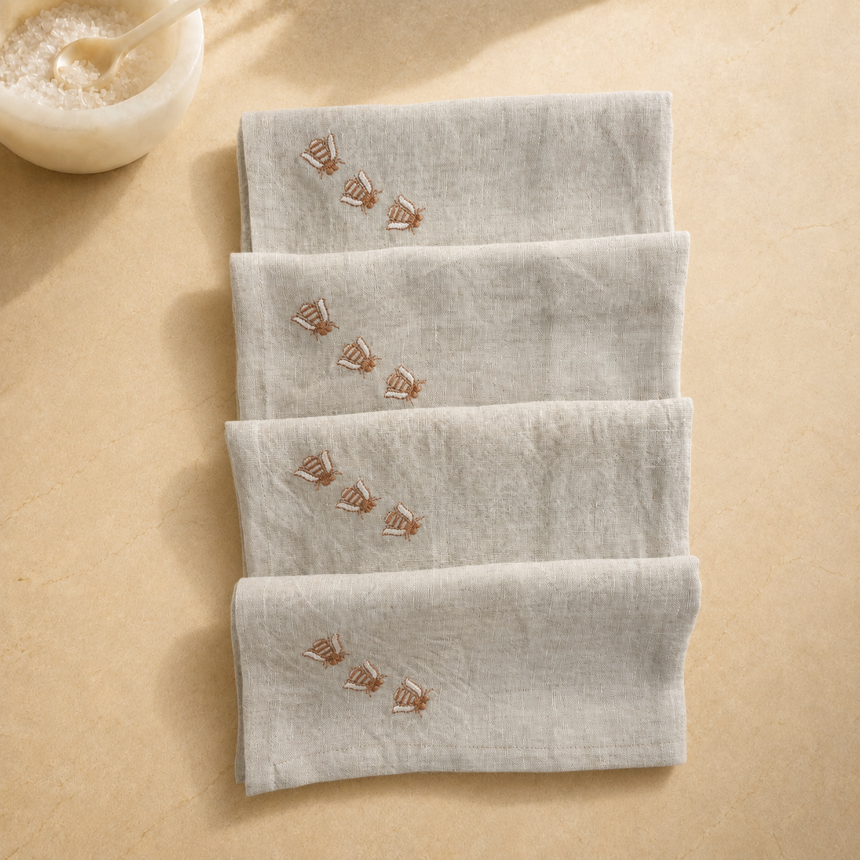 Four folded light beige linen napkins with embroidered sailboat motifs, arranged vertically on a beige surface with a small bowl in the top-left corner. MI RE LA