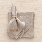 A folded light beige linen napkin with embroidered bees and a matching folded napkin underneath, tied with a twine napkin ring, on a textured beige stone surface. MI RE LA