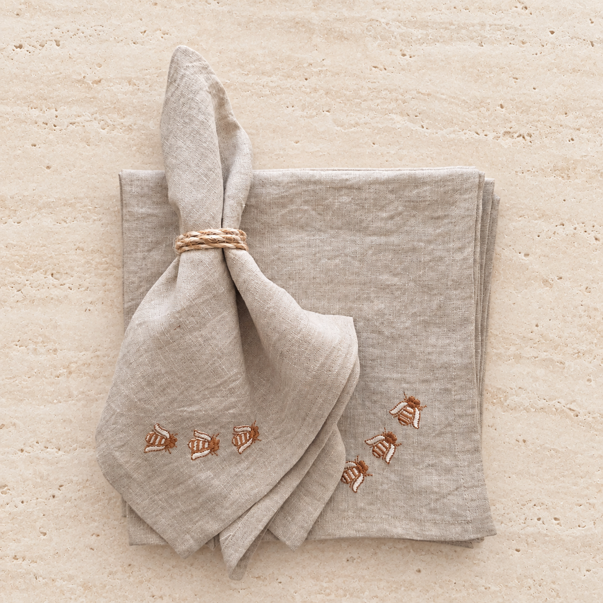A folded light beige linen napkin with embroidered bees and a matching folded napkin underneath, tied with a twine napkin ring, on a textured beige stone surface. MI RE LA