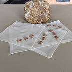 Four square white napkins with embroidered brown bees, arranged on a beige textured surface with a decorative stone behind them. MI RE LA