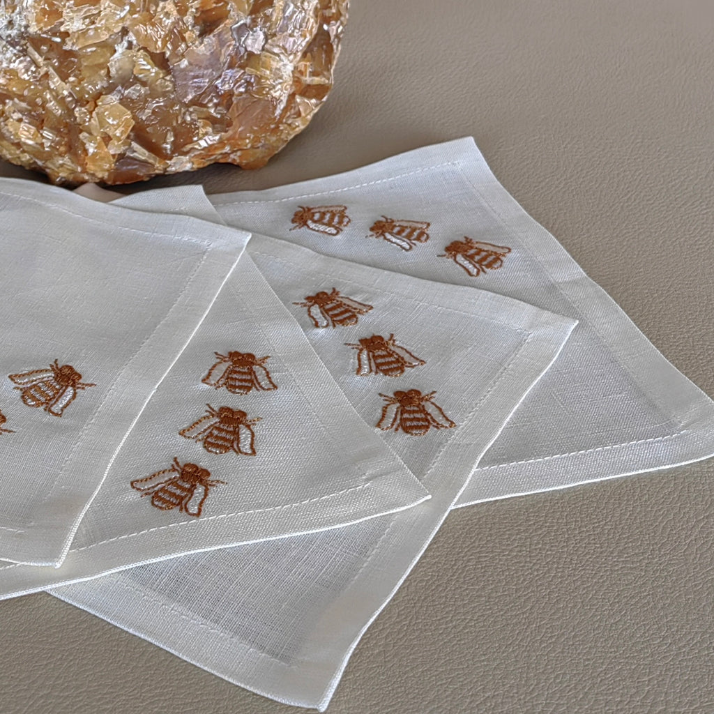 Three square white napkins with embroidered brown bees, arranged on a textured beige surface with a decorative crystal in the background. MI RE LA