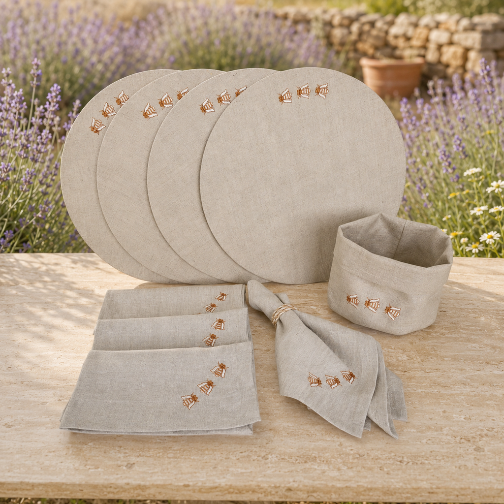 A coordinated linen table setting displayed outdoors on a wooden table, with lavender flowers and a stone wall in the background. The set includes round placemats, folded napkins, a small napkin holder, and matching embroidered cloths in a light warm gray-beige tone with small embroidered floral motifs.