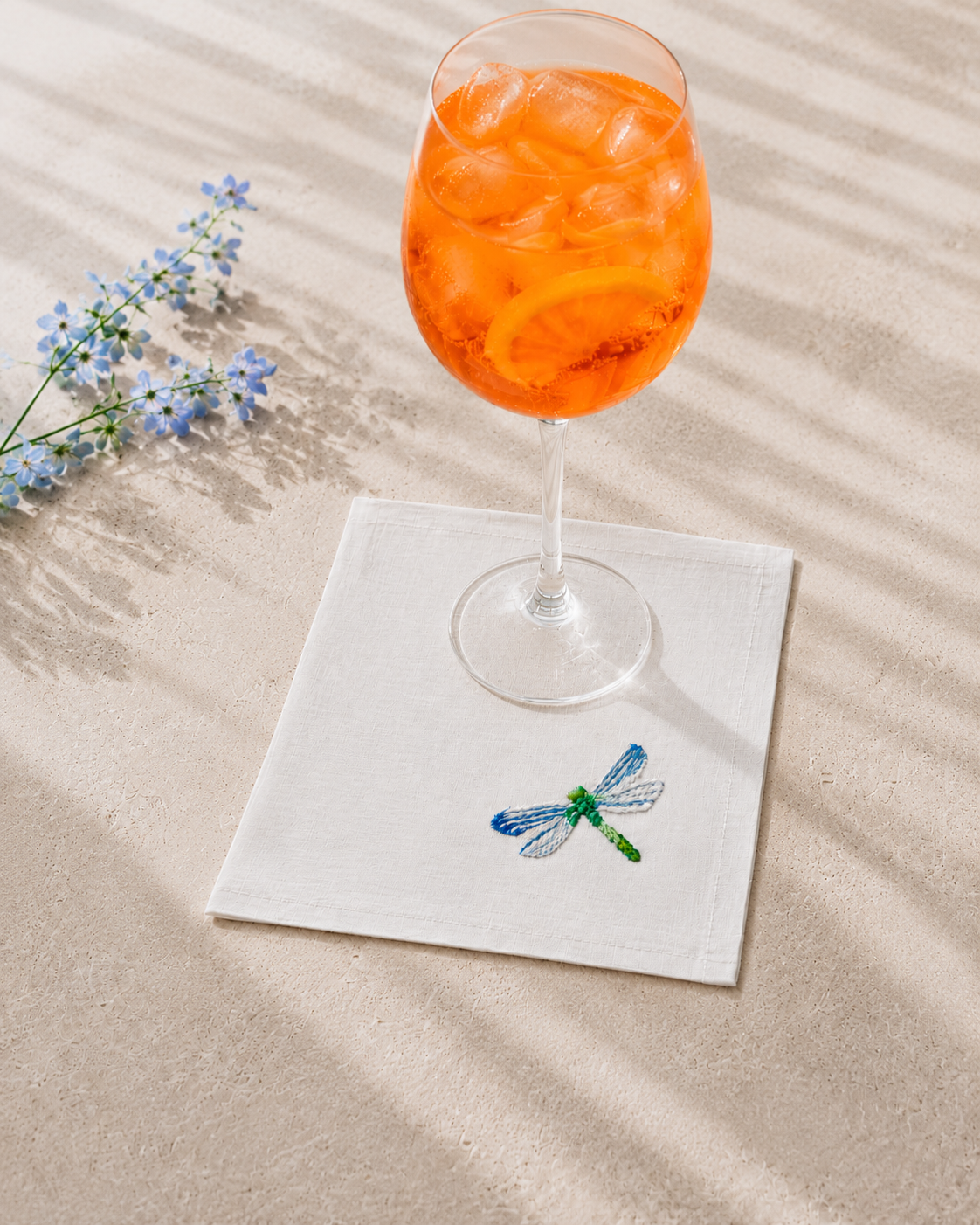 Glass of orange cocktail with ice on a white napkin with a dragonfly design, placed on sand.