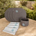A set of dark taupe rround placemat with matching gray napkins and a dark taupe basket, all featuring aqua coral embroidery, displayed on a stone table with green garden foliage in the background.