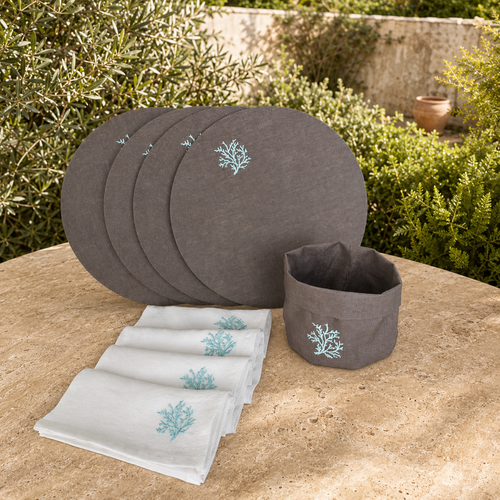 A set of dark taupe rround placemat with matching gray napkins and a dark taupe basket, all featuring aqua coral embroidery, displayed on a stone table with green garden foliage in the background.