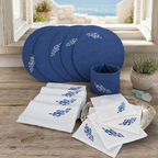 A coordinated tableware set displayed on a rustic wooden surface in front of an open window. The set includes several round blue plates, a matching blue cup, white napkins with blue embroidered motifs, and a small decorative tray.