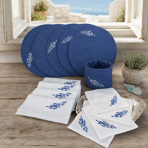 A coordinated tableware set displayed on a rustic wooden surface in front of an open window. The set includes several round blue plates, a matching blue cup, white napkins with blue embroidered motifs, and a small decorative tray.