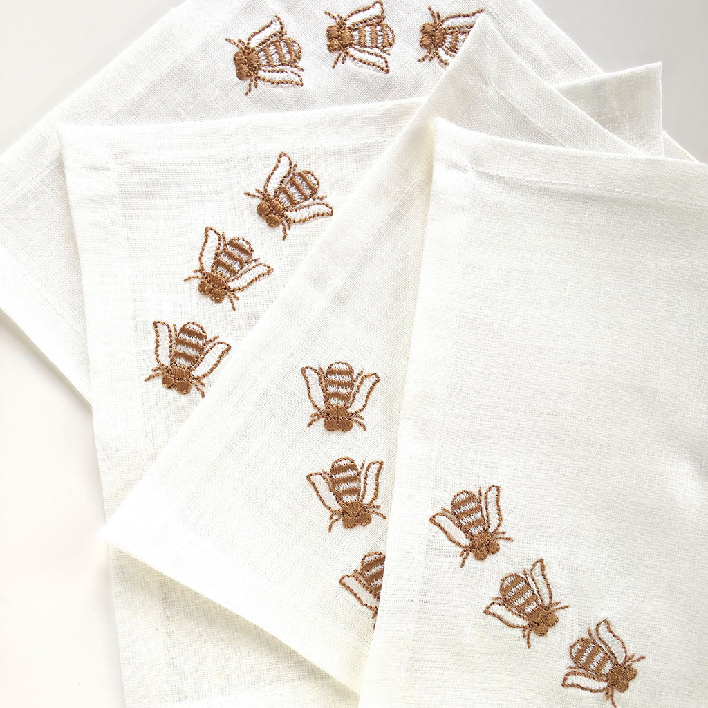 off white linen cocktail napkins with embroidered brown bees, arranged in a fan-like pattern on a white background. MI RE LA