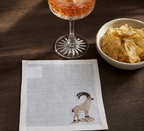 A square white linen napkin with a detailed embroidered brown and beige ram on the bottom right, styled on a dark wood table next to a glass of rosé wine and a bowl of potato chips.