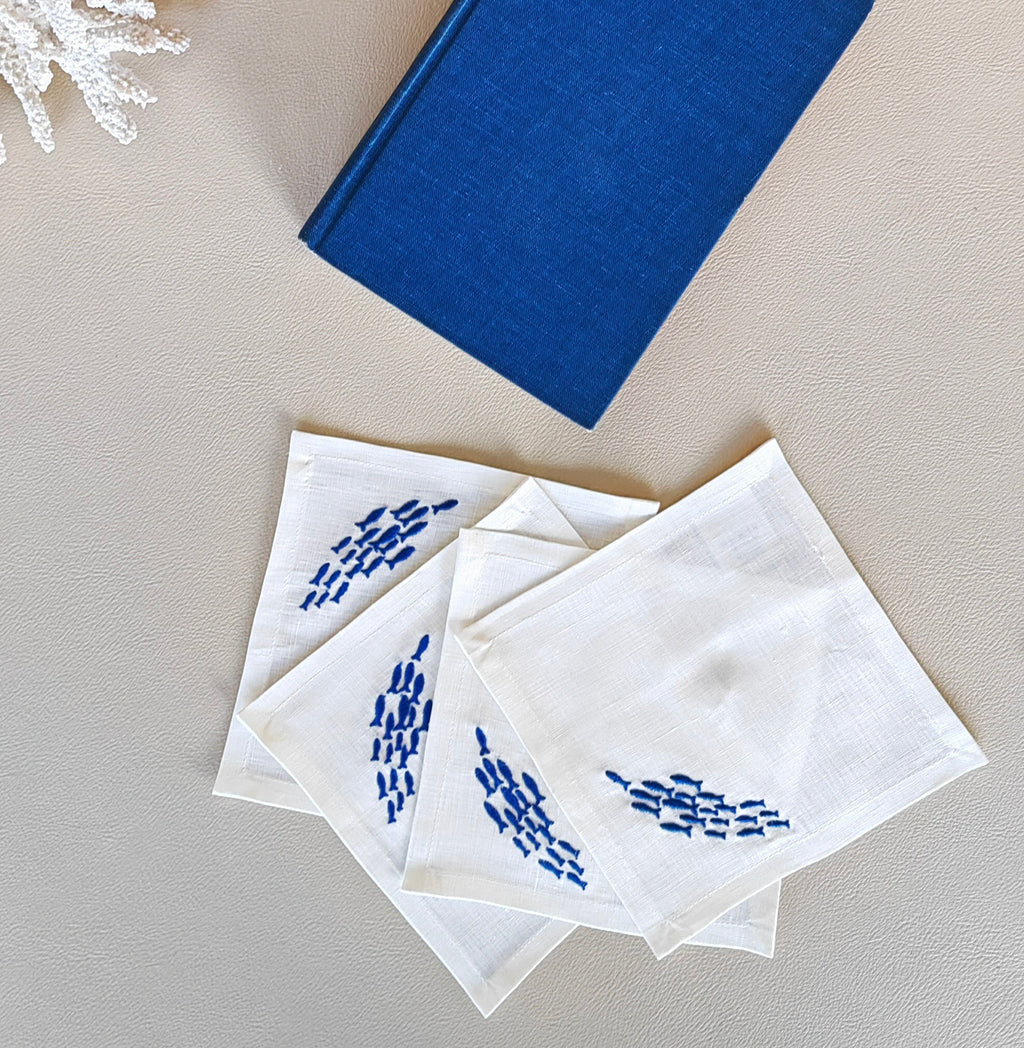 Four white square napkins with blue embroidered fish motifs, arranged on a light beige textured surface next to a blue book and a small white decorative coral.