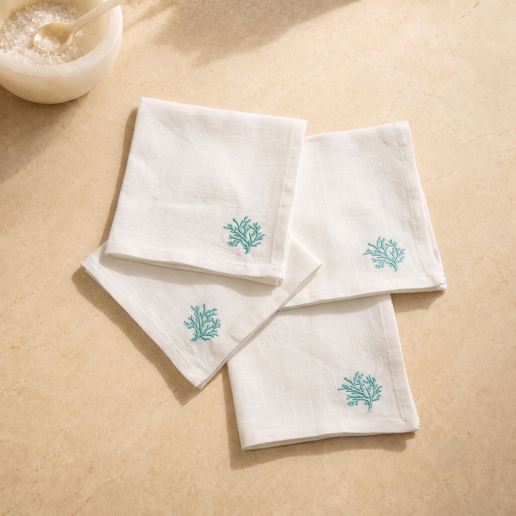 Four white Linen napkins with embroidered teal coral motifs, arranged on a beige stone surface next to a small white bowl with a spoon. MI RE LA