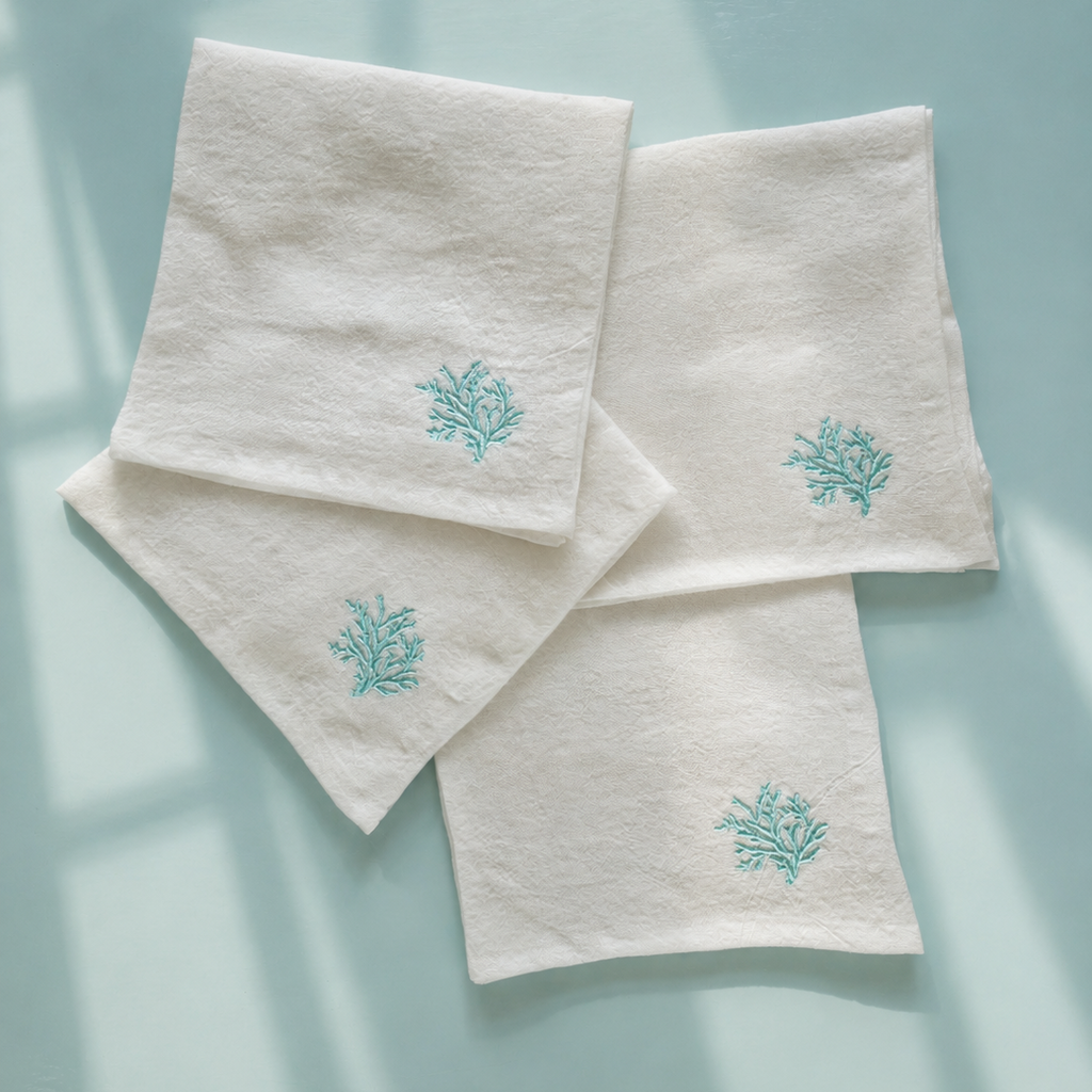 Four off-white cotton napkins with embroidered teal coral motifs, arranged in a flat-lay pattern on a light blue background with soft window shadow overlays. MI RE LA