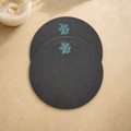 Two round dark charcoal gray coasters with light blue coral embroidery, arranged on a textured beige surface with a small white bowl in the top-left corner.