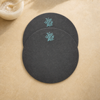 Two round dark charcoal gray coasters with light blue coral embroidery, arranged on a textured beige surface with a small white bowl in the top-left corner.