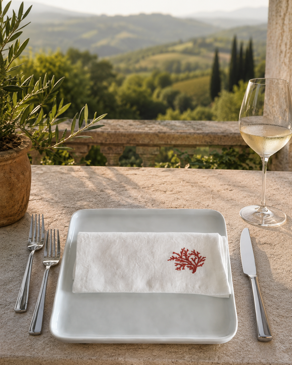 A white linen napkin with a red embroidered coral motif, folded on a square white plate on a stone table. Silver forks and a knife are placed on either side, with a glass of white wine and a potted olive plant in the foreground, set against a scenic Tuscan vineyard landscape. MI RE LA