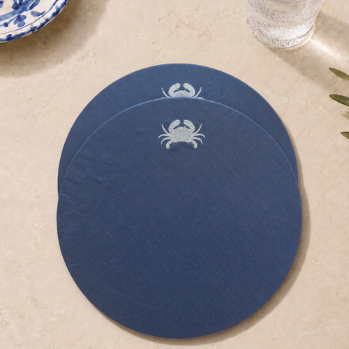 Two round navy blue coasters stacked on a beige stone surface, each printed with a small white crab motif near the top edge. MI RE LA