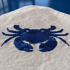 Close-up of a beige woven textile with a prominent embroidered blue crab motif, set against a dark blue background. MI RE LA