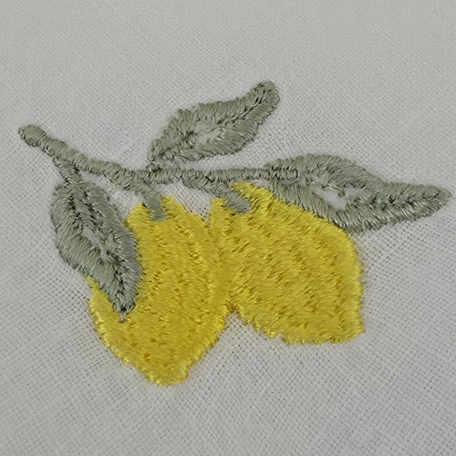 A close-up of embroidered lemons on a white fabric background. The embroidery features two bright yellow lemons with textured stitching and gray-green leaves and stems. MI RE LA