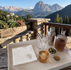 A styled outdoor bar setup on a wooden balcony with a dramatic mountain landscape in the background. The tray holds a folded white napkin with a floral print, a glass of amber drink with an ice cube and orange peel, a shaker, a bottle, olives, and a small dish of nuts.