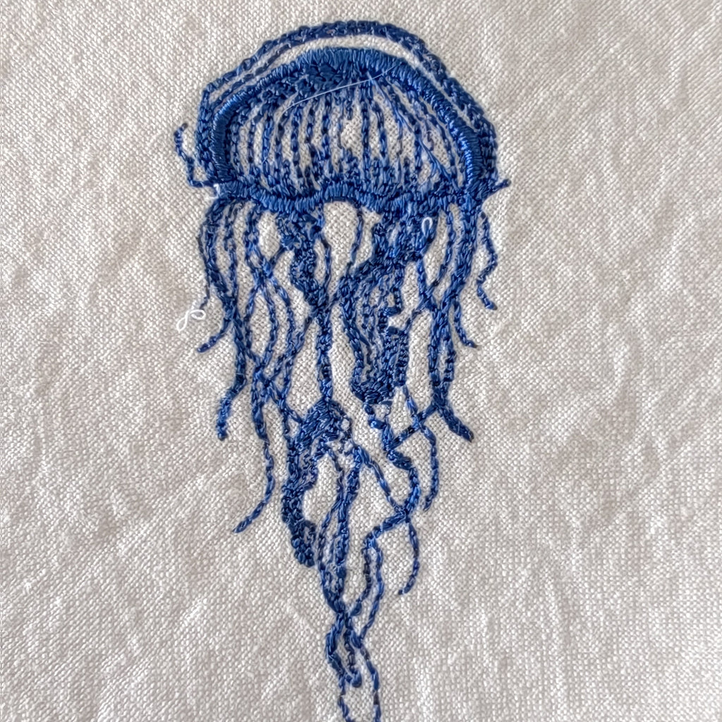 A blue embroidered jellyfish design on a textured off-white fabric background.