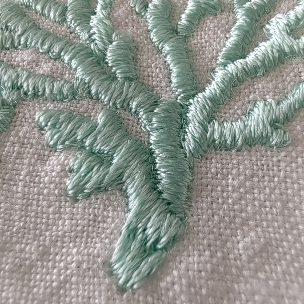 Close-up of mint green embroidered branches or coral-like stitching on a light beige woven fabric background. MI RE LA