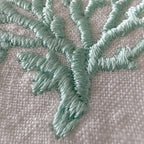 Close-up of mint green embroidered branches or coral-like stitching on a light beige woven fabric background. MI RE LA