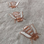 Close-up of a light gray textured fabric with three embroidered butterflies in dusty rose and white thread. MI RE LA